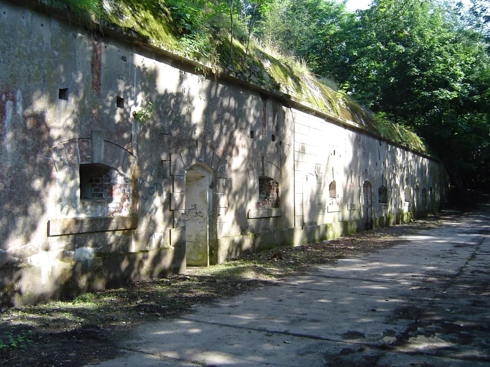 A long, weathered concrete wall with small arched openings and patches of moss, under the shade of trees.